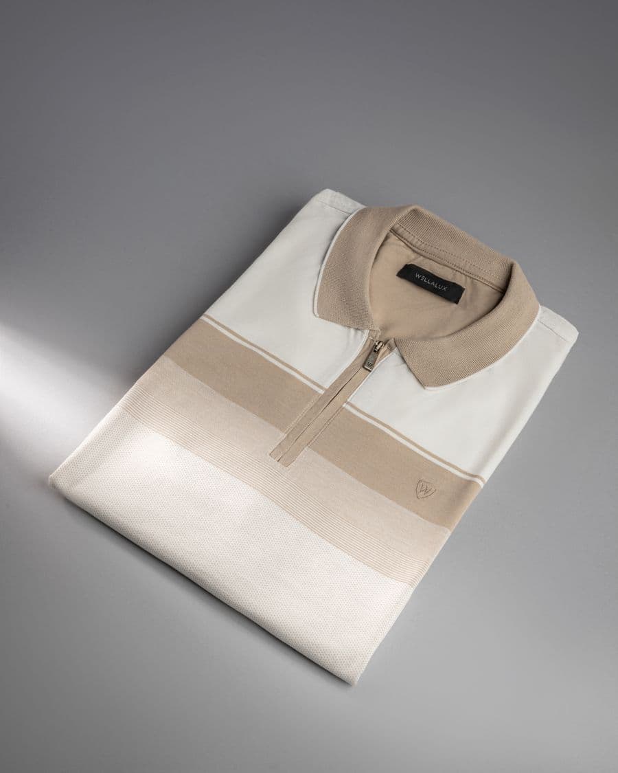 A folded zip-neck polo shirt in cream and sand panels, photographed flat on a plain grey surface.