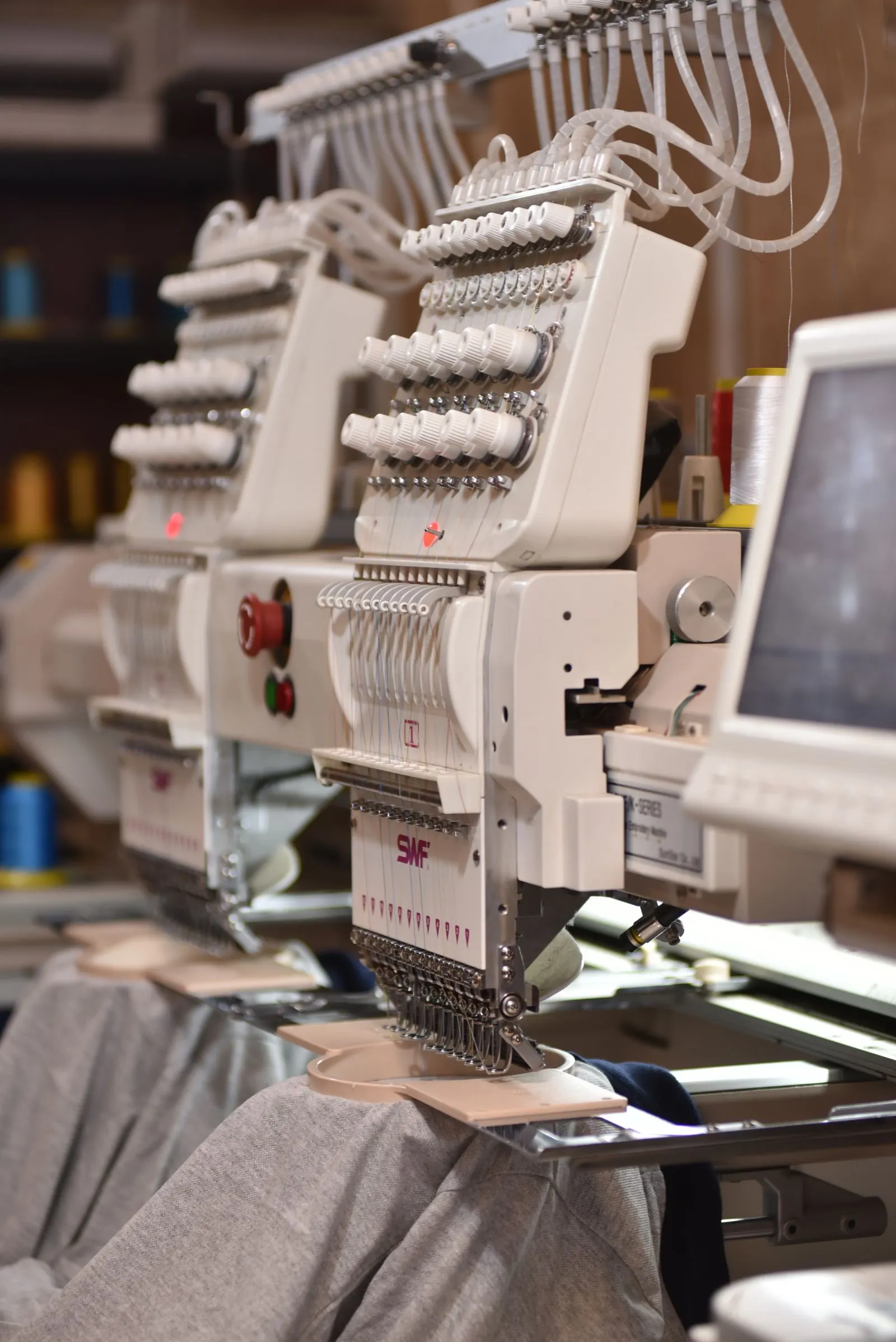 Two multi-head embroidery machines on a workshop floor, threaded and mid-run, with a grey garment hooped under the needles and thread cones racked behind.