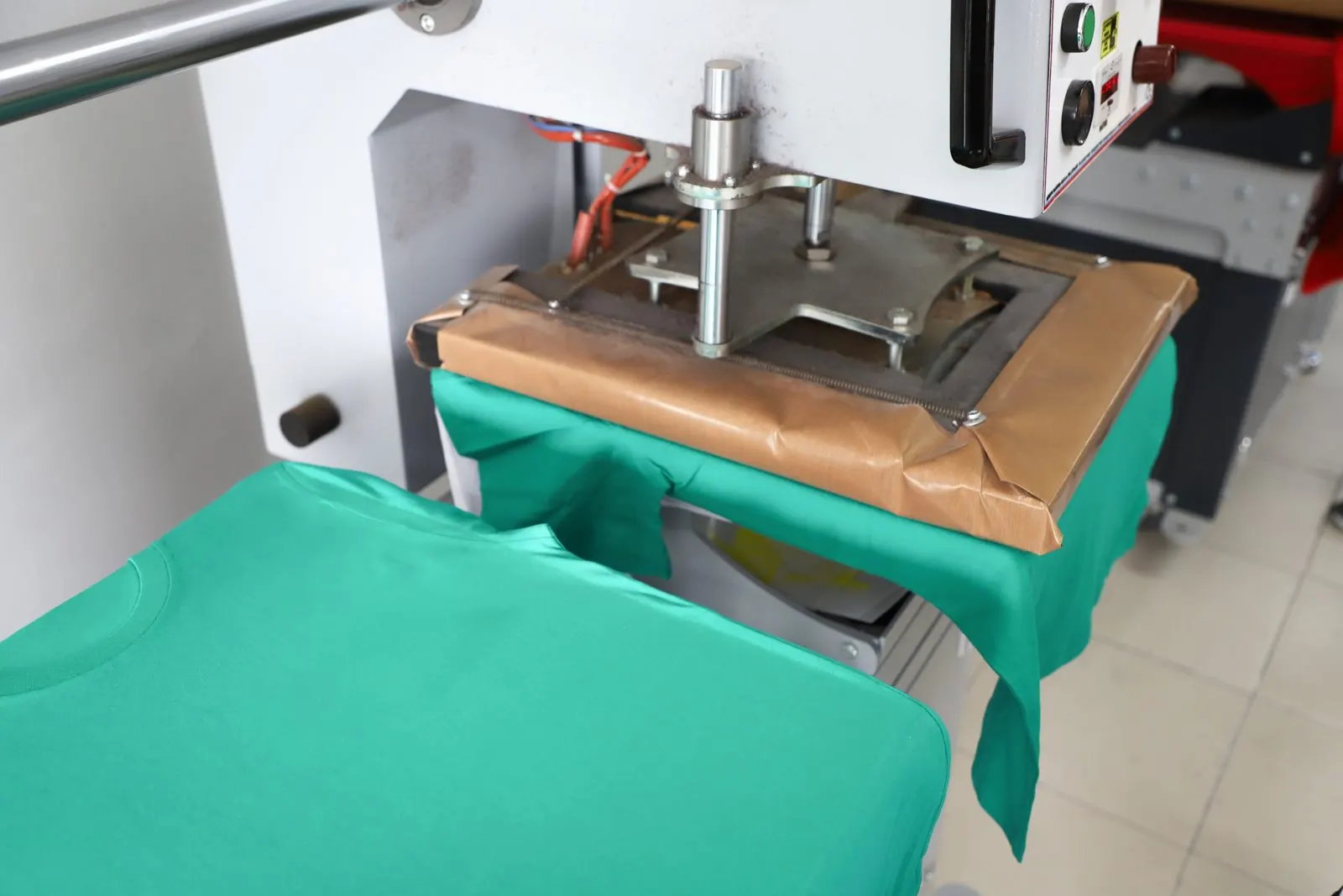 A heat press mid-cycle on a print floor, platen lowered onto a green T-shirt with transfer paper taped across the print area.